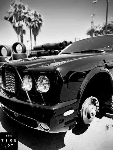 A black luxury sedan on a jack with the front wheel removed and the brake rotor exposed, tire racks and palm trees behind it.