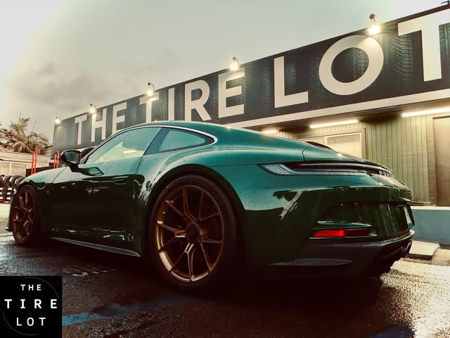 A dark green sports car parked at dusk under the lit THE TIRE LOT sign on the front of the shop.