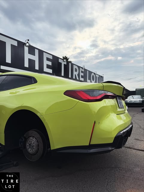 A bright yellow-green coupe raised on a jack with a wheel removed, under the THE TIRE LOT sign.
