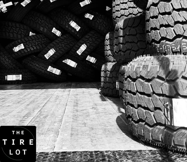 Black and white view inside the shop: a wall of stacked tires with size labels, next to oversized truck tires on a concrete floor.